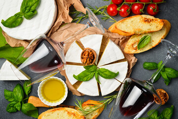 Cheese and wine background. Brie cheese, glasses of wine, nuts and grapes on a table with basil, olive oil and toasted bread. On a stone background. Top view.
