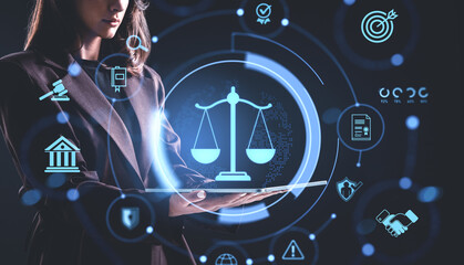 Woman holding tablet with glowing justice scale hologram surrounded by legal icons on dark...
