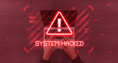 Cybersecurity alert with red warning sign and text "System Hacked" over hands typing on laptop with digital glitch effect on wood background.