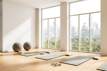 Bright yoga studio interior design with large windows, scenic city view, exercise mats, dumbbells, and fitness balls on wooden floor. 3D Rendering