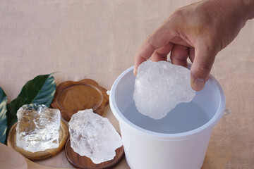Close up hand holds large clear alum crystal over a white container with more crystal pieces placed on wooden trays. Concept of natural minerals, purification, traditional use, and chemical compounds