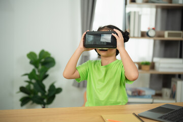 Child Engaged in Virtual Reality Experience with VR Headset while Sitting at Table in Modern Room with Laptop and Indoor Plant in Background