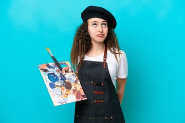 Young artist Arab woman holding a palette isolated on blue background and looking up