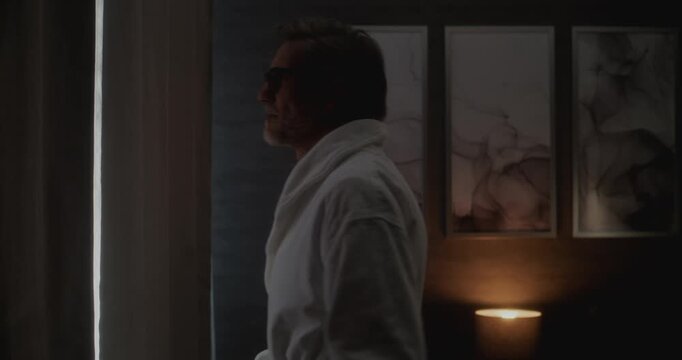 Mature Man in Soft White Robe Opens Curtains in Cozy Hotel Room, Letting in Warm Sunshine. Peaceful Smile Reflects Calm Start to Day. Concept of Mindful Travel, Morning Balance, and Inner Harmony.