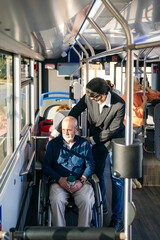 Man in a sharp suit assisting a senior man in a wheelchair on a city bus, promoting accessibility for elderly passengers