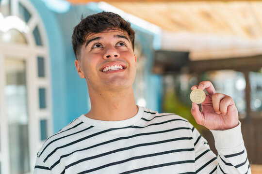 Young hispanic man holding a Bitcoin at outdoors looking up while smiling