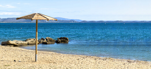 A wooden beach umbrella stands upon the quiet sandy shore beside clear blue sea and scattered rocks under bright daylight, featuring distant hills visible across the calm horizon