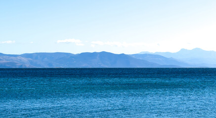 Tranquil blue sea stretching toward the distant mountain horizon under clear midday light, captured from a low coastal viewpoint featuring gentle ripples and soft atmospheric tones