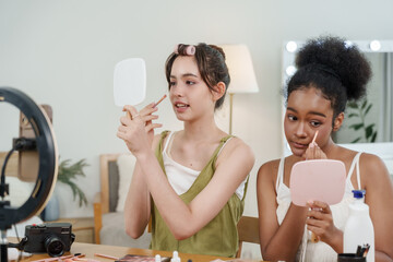 asian teenage beauty vlogger and black teenage friend applying makeup in front of mirror smiling enjoying beauty routine sitting together at table filled with skincare cosmetics content creation tools