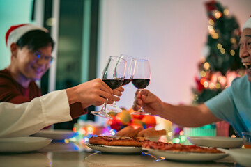 Asian family celebrating Christmas Eve at home, toasting red wine around the dinner table filled with pizza and holiday cheer.