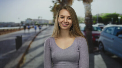 Fototapeta premium Woman smiling outdoors on a sunny beach promenade with parked cars and blurred sea, showcasing casual elegance in an outdoor setting.