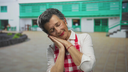 Senior woman with grey hair in apron making sleep gesture on an urban street in an old town...