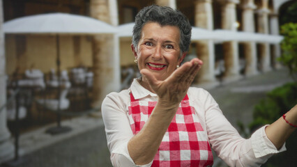 Obraz premium Senior woman with grey hair and apron joyfully extends hands in an inviting gesture on a charming old town street with outdoor cafes and arches.