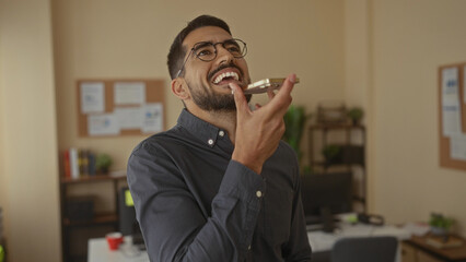 Young man using smartphone in modern office setting, displaying cheerful expression with plants in background indicating a casual professional environment.