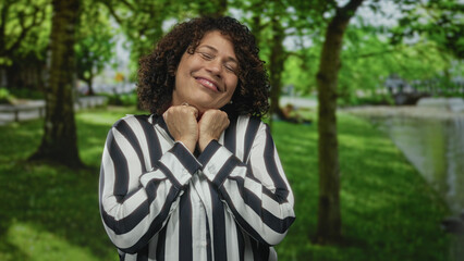 Middle aged woman wearing striped blouse crosses index fingers in lush forest park under bright sunlight; defiance.