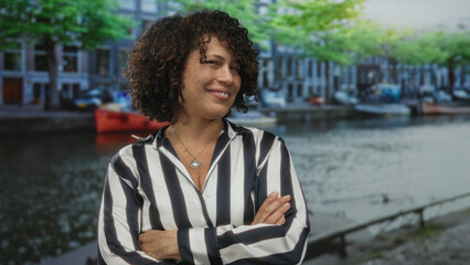 Woman crosses arms on canal street in amsterdam by historic building facade while smiling at the...