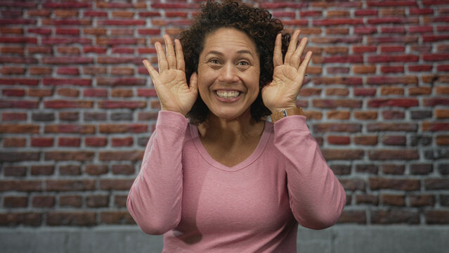 Woman sticking out her tongue and removing hands from her face in a brick wall studio setting, wearing pink sweater and wooden watch; playful.