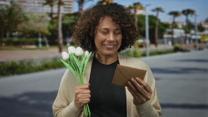 Woman smiling while holding a white tulip bouquet and a brown envelope on an urban street;...