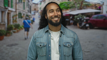 Fototapeta premium Man smiling with visible face and beard on a cobblestone street near cafe tables and scooters; happiness optimism.