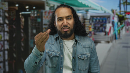 Fototapeta premium Man making hand gesture in busy street market among souvenir stalls and storefronts; frustration complaint.