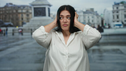 Woman in white shirt stands in vatican city square covering ears with a calm expression, portraying a peaceful moment amid bustling outdoor urban scene.