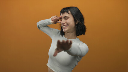 Fototapeta premium Smiling young hispanic woman making peace sign near eye in studio against orange wall; serenity joy.