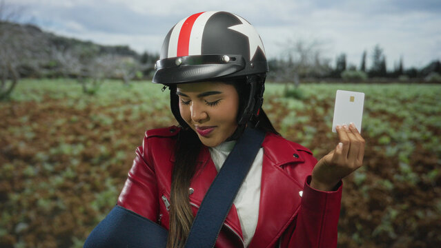Young woman in helmet and red jacket with arm in sling holding credit card outdoors in green park setting.
