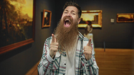 Young bearded man wearing plaid shirt points finger to painting on wall in building with wide open mouth; excitement.
