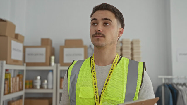 Young hispanic man in reflective vest volunteering at an indoor donations center gesturing while holding a clipboard amid organized shelves and boxes.