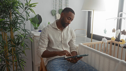 Man taps tablet beside baby crib in a nursery room, hand touching screen while seated on chair by window; calm focus.