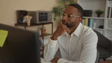 Fototapeta premium Man wearing white shirt and round glasses rests chin on hand at office desk while studying a monitor; pensive concentration.
