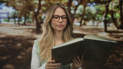 Obraz premium Blonde woman holds open hardcover book with both hands reading under tall oak trees in green city park; calm reflection.