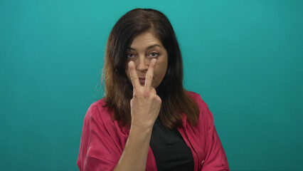 Middle aged hispanic woman makes hand binoculars gesture to face in a studio with teal backdrop;...