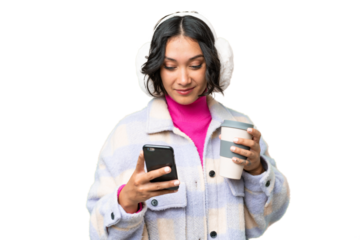 Young Argentinian woman wearing winter muffs over isolated background holding coffee to take away and a mobile