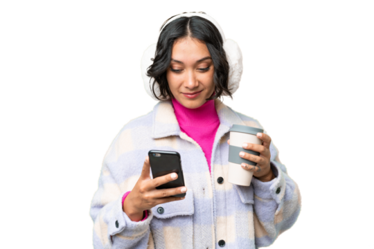 Young Argentinian woman wearing winter muffs over isolated background holding coffee to take away and a mobile