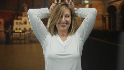 Woman holds horns gesture with fingers amid pew and altar details in church while smiling broadly;...