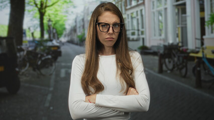Fototapeta premium Woman with arms crossed on a city street wearing glasses and a white top facing camera with stern expression; annoyed defiance.