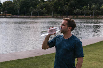 Man drinking refreshing water from a plastic bottle, staying hydrated outdoors beside a tranquil...