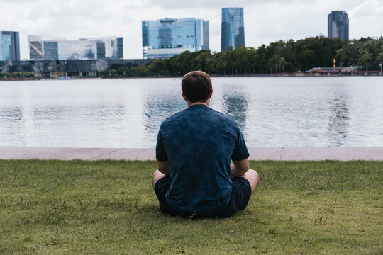 Man sitting on grass by a city lake, looking at the distant urban skyline, representing contemplation and tranquility