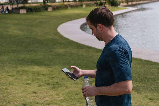 Adult man standing near a lake in park, checking his smartphone and holding a water bottle, taking a break outdoors