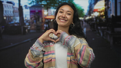 Fototapeta premium Woman makes heart with hands and points forward on a busy city street while smiling at camera and showing bare hands; joy affection.