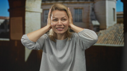 Fototapeta premium Blonde woman covers ears against sunlit facade of historic building on narrow old town street; anxiety.