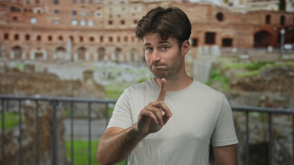 Young man points finger forward near roman ruins building, standing by metal railing with a wry...