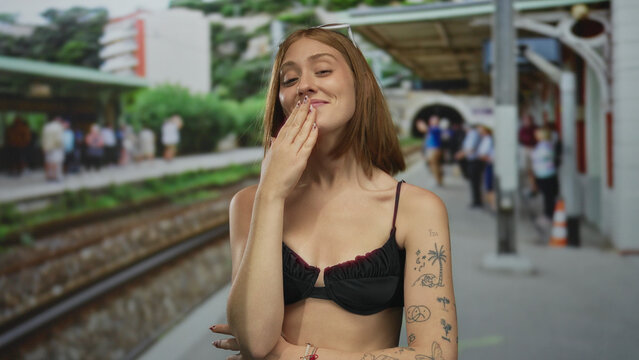 Young woman with tattoos smiling at an outdoor train station, surrounded by people waiting near the railway tracks during a sunny day.