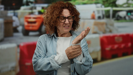 Woman examining hand at a busy construction site, showcasing concern, with orange machinery and red barriers in the background, highlighting the outdoor work scenario.