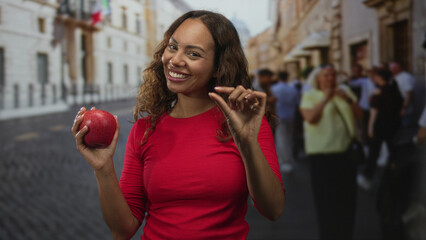Woman smiling and holding a red apple in her left hand while pinching fingertips with her right on a cobblestone street; playful.