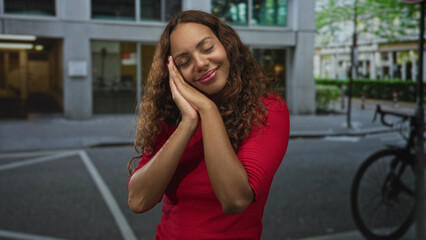 Fototapeta premium Young woman wearing a red top with hands pressed to cheek in a sleepy gesture on a street by a parked bicycle; serenity rest.