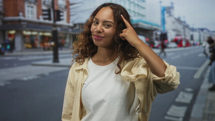 Obraz premium Woman points finger to temple on urban city street, smiling at camera while wearing white t shirt and beige overshirt, curly hair framing face; confidence.