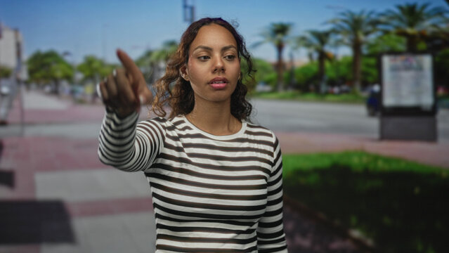 Woman points finger up on street, visible hand and face in striped top with index finger extended, palm forward; determination ambition.