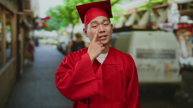 Young man wearing red graduation gown and cap stands on a bustling city street displaying a thoughtful expression, capturing a moment of post-graduation reflection outdoors.
